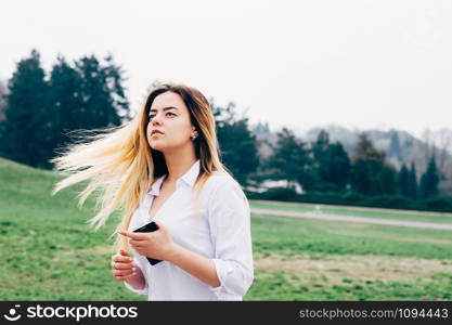 A portrait of a pretty young long haired woman in white shirt, standing in the windy park and looking into the distance, holding a cellphone in her hand