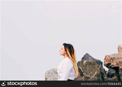 A portrait of a pretty young long haired woman in white shirt, standing on a beach or hill, with big stones in the background and much air for copy space. Romantic or freedom concept