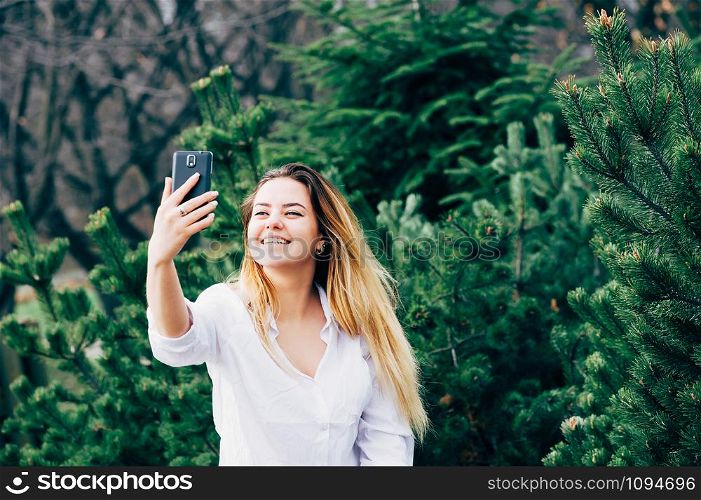 A portrait of a pretty young long haired woman in white shirt, smiling and making selfie with her smartphone in a park, with green pine trees in the background