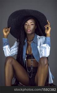 A portrait of a seductive young black female with long dreadlocks, beautiful makeup, moist lips posing by herself in a studio with grey background wearing a summer hat & outfit with jewelry.