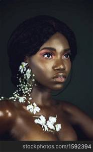 A portrait of a sensual young black female with curly long black hair, beautiful makeup and moist lips posing by herself in a studio with dark background wearing white flowers on her ears & chest.