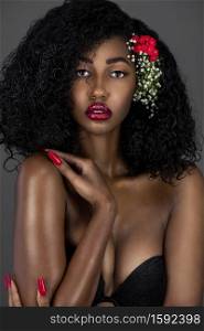 A portrait of a sensual young black female with curly long black hair, beautiful makeup and red lips posing by herself in a studio with grey background wearing flowers in her hair.