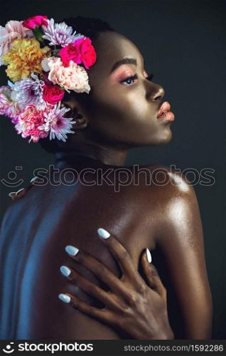 A portrait of a serene young black female with a short Afro, beautiful makeup and moist lips posing by herself in a studio with dark background wearing a bouquet of flowers in her hair.