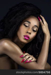 A portrait of a serene young black female with long black curly hair, beautiful makeup, fuchsia lips and nails posing by herself in a studio with a dark background.