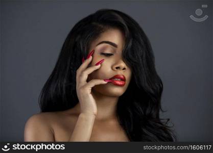 A portrait of a serene young Latina with long black wavy hair, beautiful makeup, red lips and nails posing by herself in a studio with a grey background.