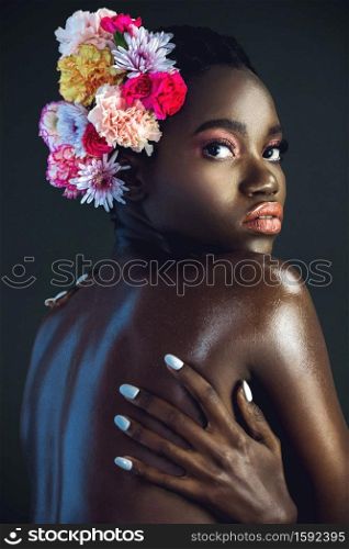 A portrait of a sexy young black female with a short Afro, beautiful makeup and moist lips posing by herself in a studio with dark background wearing a bouquet of flowers in her hair.