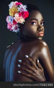 A portrait of a sexy young black female with a short Afro, beautiful makeup and moist lips posing by herself in a studio with dark background wearing a bouquet of flowers in her hair.