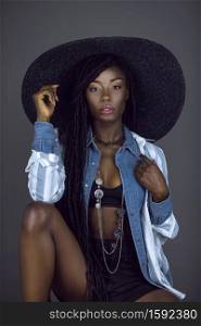 A portrait of a sexy young black female with long dreadlocks, beautiful makeup, moist lips posing by herself in a studio with grey background wearing a summer hat & outfit with jewelry.