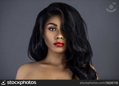 A portrait of a sexy young Latina with long black curly hair, beautiful makeup and red lip stick posing by herself in a studio with a grey background.