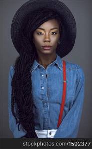 A portrait of a young black female with long dreadlocks and beautiful makeup posing by herself in a studio with grey background wearing a summer hat, denim shirt & red suspenders.