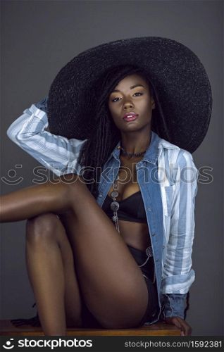 A portrait of a young black female with long dreadlocks, beautiful makeup, moist lips posing by herself in a studio with grey background wearing a summer hat & outfit with jewelry.