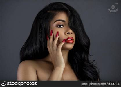 A portrait of a young Latina with long black wavy hair, beautiful makeup, red lips and nails posing by herself in a studio with a grey background.