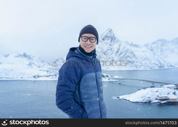 A Portrait of Asian tourist man standing on hill and traveling in Lofoten islands, Norway, Europe. White snowy mountain hills, nature landscape in winter. Trekking and hiking. Adventure.