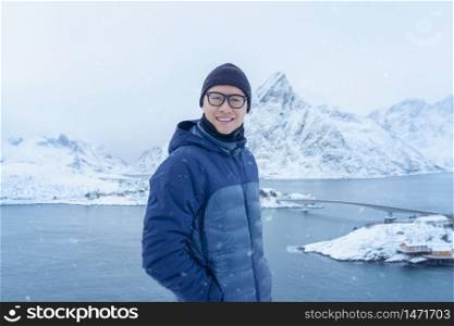 A Portrait of Asian tourist man standing on hill and traveling in Lofoten islands, Norway, Europe. White snowy mountain hills, nature landscape in winter. Trekking and hiking. Adventure.
