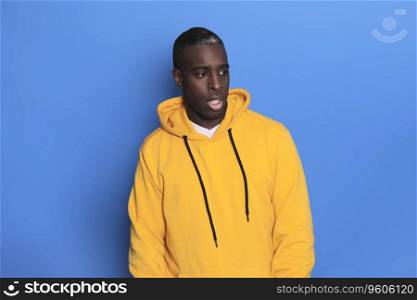 A Portrait of cheerful young african-american guy wearing yellow hoodie sweater posing isolated on blue background. Carefree young millennial man looking aside