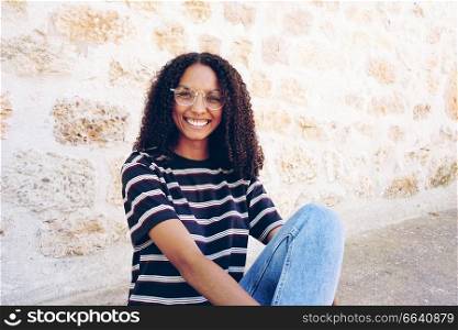 A portrait of happy smiling young black woman wearing glasses, jeans and a striped t-shirt, sitting on the ground and curly hair