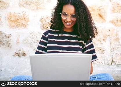A portrait of happy smiling young black woman with curly hair wearing glasses, jeans and a striped t-shirt, sitting on the ground and working or making homework