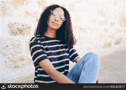 A portrait of serious young black woman wearing glasses, jeans and a striped t-shirt , sitting on the ground and curly hair