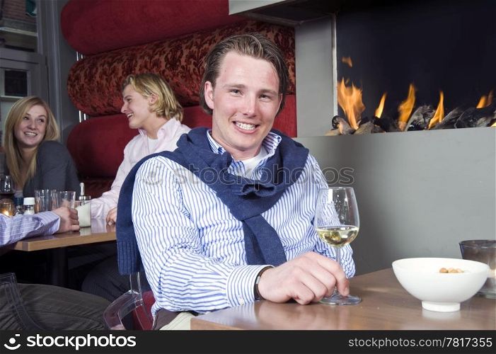 A posh looking man with his sweater around his neck and a glass of white wine in his hand sitting at a restaurant table in front of the fireplace