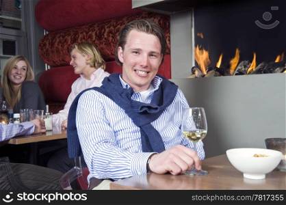 A posh looking man with his sweater around his neck and a glass of white wine in his hand sitting at a restaurant table in front of the fireplace