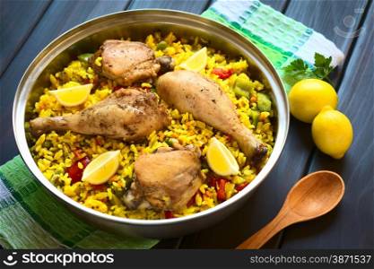 A pot of chicken paella, a traditional Valencian (Spanish) rice dish made of rice, chicken, peas, capsicum and served with lemon, photographed on dark wood with natural light (Selective Focus, Focus on the middle of the dish)