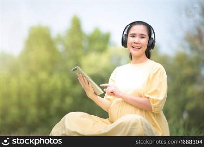 A pregnant woman is relaxing in the park and listening to music on a tablet, Mom listening Songs for the unborn child, Pregnant near giving birth, mother and child