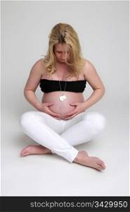 A pregnant young women sitting down embracing her belly and watching towards it. The women is wearing a black top and a white chino on bare feet