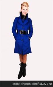 A pretty and tall woman in a blue winter coat and black boots standing inthe studio, her hands in the pocket and legs crossed, for white background.