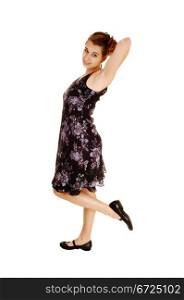 A pretty and young woman in a black dress with flowers on standing fromthe side for white background, lifting up one leg.