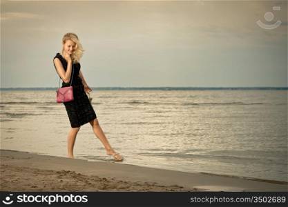 A pretty blonde in a black dress standing barefoot on the beach.