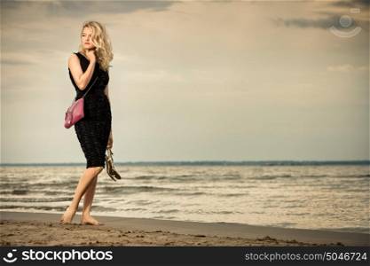 A pretty blonde in a black dress standing barefoot on the beach.