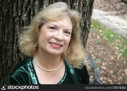A pretty, fifty year old blond woman leaning against a tree in the park.