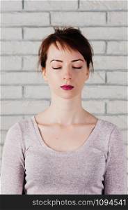 A pretty girl with eyes closed as in meditation, peaceful look, with white brick wall in the background. Face mimic for fitness exercise