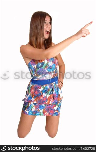 A pretty teenage girl kneeling on the floor in a colorful dress and pointingwith her finger, with long dark blond hair, for white background.