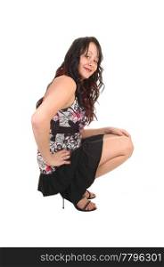 A pretty woman crouching on the floor in a short black skirt and with longcurly black hair, smiling inti the camera for whiye background.