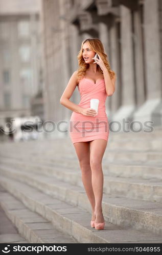 A pretty woman in a pink dress talking via mobile phone and holding a coffee cup.