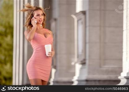 A pretty woman in a pink dress talking via mobile phone and holding a coffee cup.