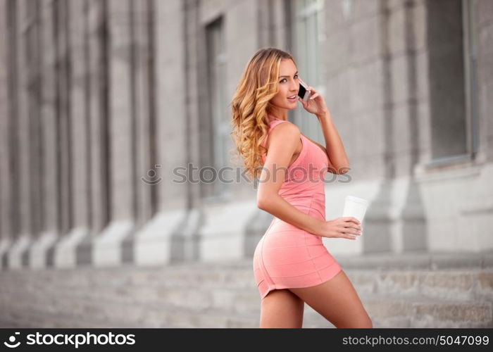 A pretty woman in a pink dress using mobile phone and holding a take away coffee.