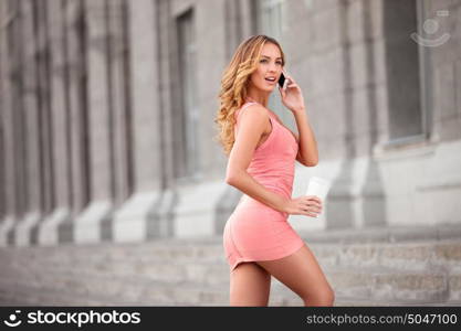 A pretty woman in a pink dress using mobile phone and holding a take away coffee.