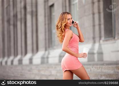 A pretty woman in a pink dress using mobile phone and holding a take away coffee.