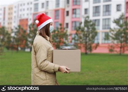 a pretty woman in a red Santa Claus hat and medical protective mask holds a big box outdoor. New Year&rsquo;s Christmas parcel in box, delivery online store in Quarantine time. Service coronavirus. a pretty woman in a red Santa Claus hat and medical protective mask holds a big box outdoor. New Year&rsquo;s Christmas parcel in box, delivery online store in Quarantine time. Service coronavirus.