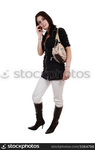 A pretty woman in white pants and brown boots in a black jacket, standing in the studio on her phone with her purse over her shoulder,for white background.
