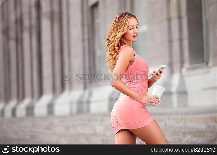 A pretty woman with a mobile phone holding a take away coffee against urban scene.