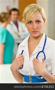 A pretty young nurse with a stethoscope shot with more medical staff out of focus behind her.