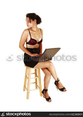 A pretty, young woman with a laptop on her lap, sitting on a chair in ablack skirt and black red bra with legs crossed for white background,looking back.