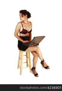 A pretty, young woman with a laptop on her lap, sitting on a chair in ablack skirt and black red bra with legs crossed for white background,looking back.