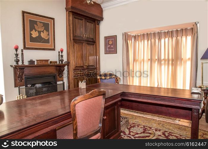 A private study in a residential home with wooden stydy table, chair, fireplace and cupboard in view near the winow.