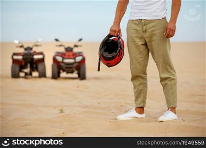A racer holds helmet near atvs, freedom riding in desert. Male persons on quad bikes, sandy race, dune safari in hot sunny day, 4x4 extreme adventure, quad-biking. A racer holds helmet near atvs in desert