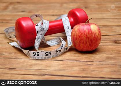 A Red dumbbell on a wooden background with a measuring tape and an apple