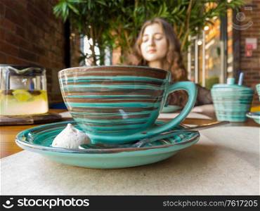 A red-haired girl sits at a table in a cafe blurry image shot, in the foreground a ceramic cup of green color with a pattern. A red-haired girl sits at a table in a cafe blurry image, in the foreground of a ceramic cup